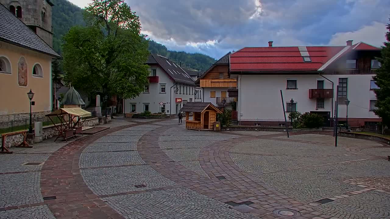 A cobblestone town square is surrounded by buildings, including a church and houses, under a cloudy sky.