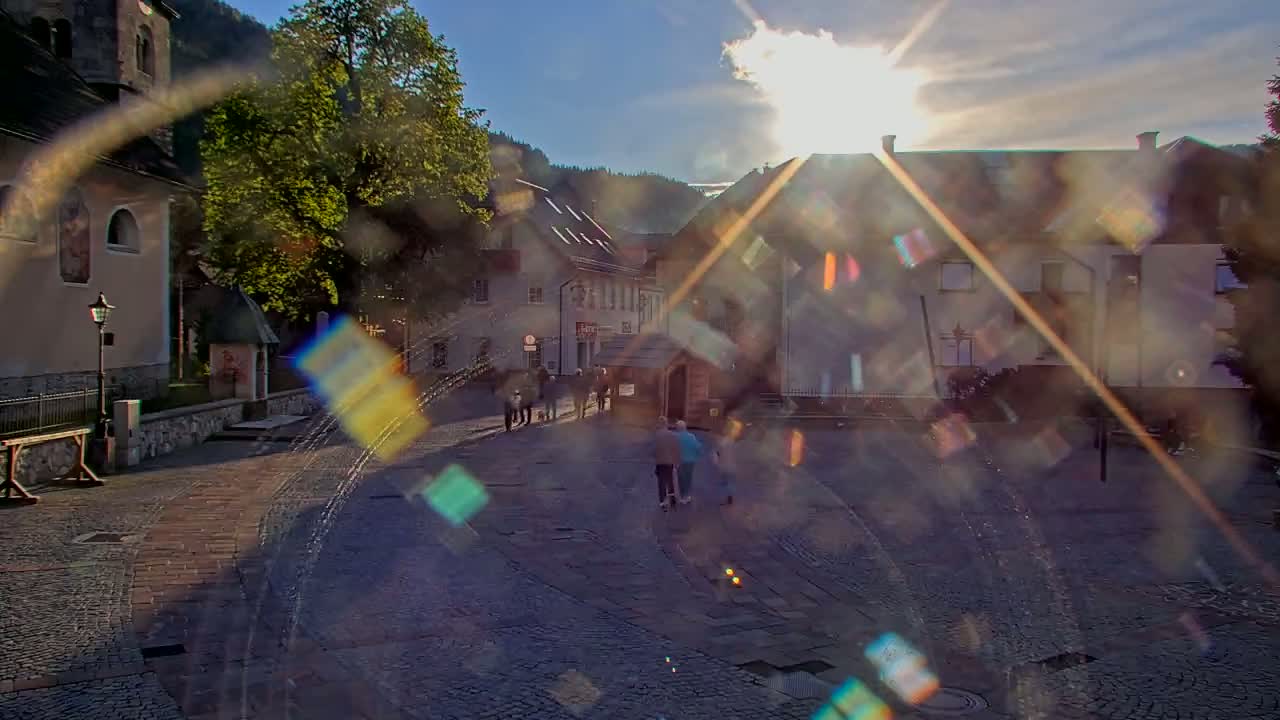 A sun-drenched cobblestone town square, surrounded by buildings and trees, with people walking on a bright, clear day.