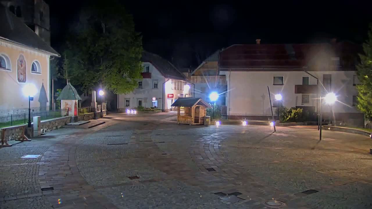 A cobblestone town square at night is illuminated by lampposts, with buildings and trees lining its perimeter.