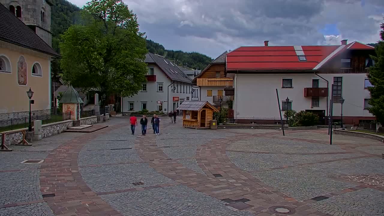 A cobblestone town square, under a cloudy sky, is surrounded by buildings, including a church and several multi-story structures, with a few people walking.