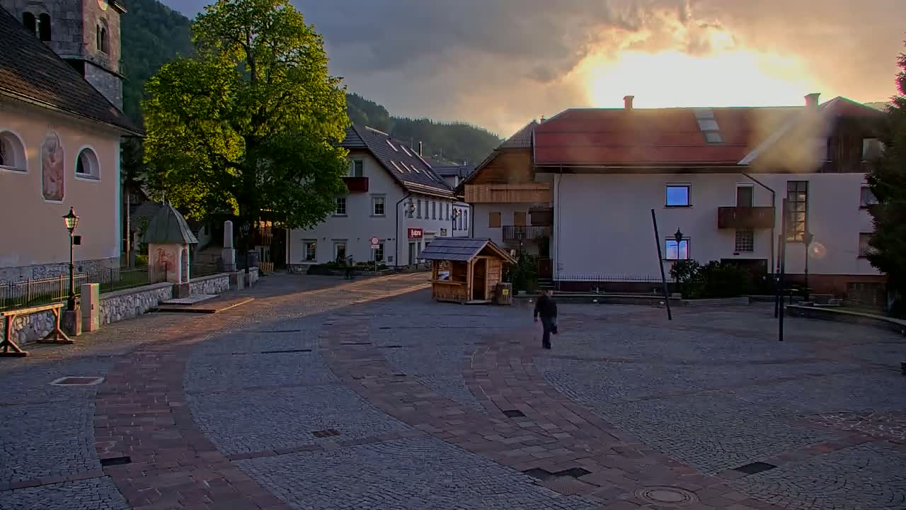 A person walks across a cobblestone town square, surrounded by buildings and a large tree, under a partly cloudy sunset.