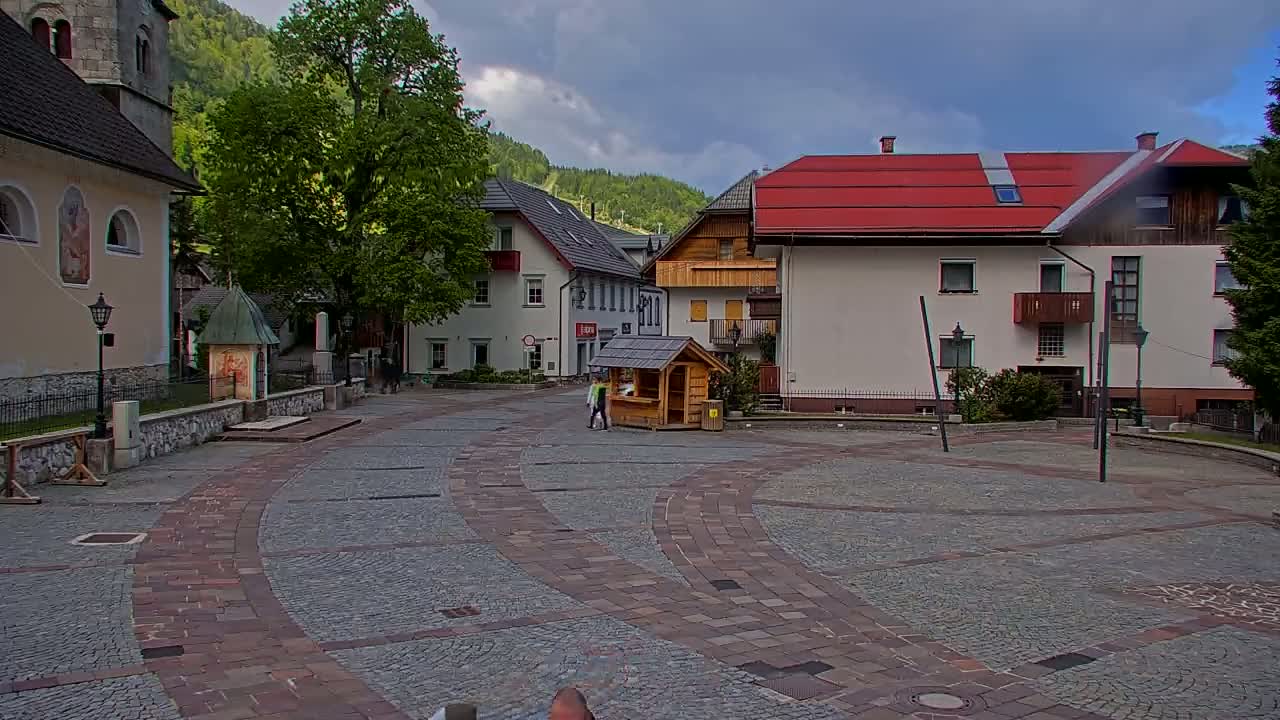 A cobblestone town square, under a partly cloudy sky, is surrounded by buildings, including a church and several multi-story houses, with a small wooden kiosk in the center.