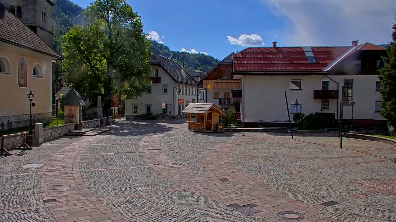 A cobblestone town square, under a partly sunny sky, is surrounded by European-style buildings and a lush green mountain backdrop.