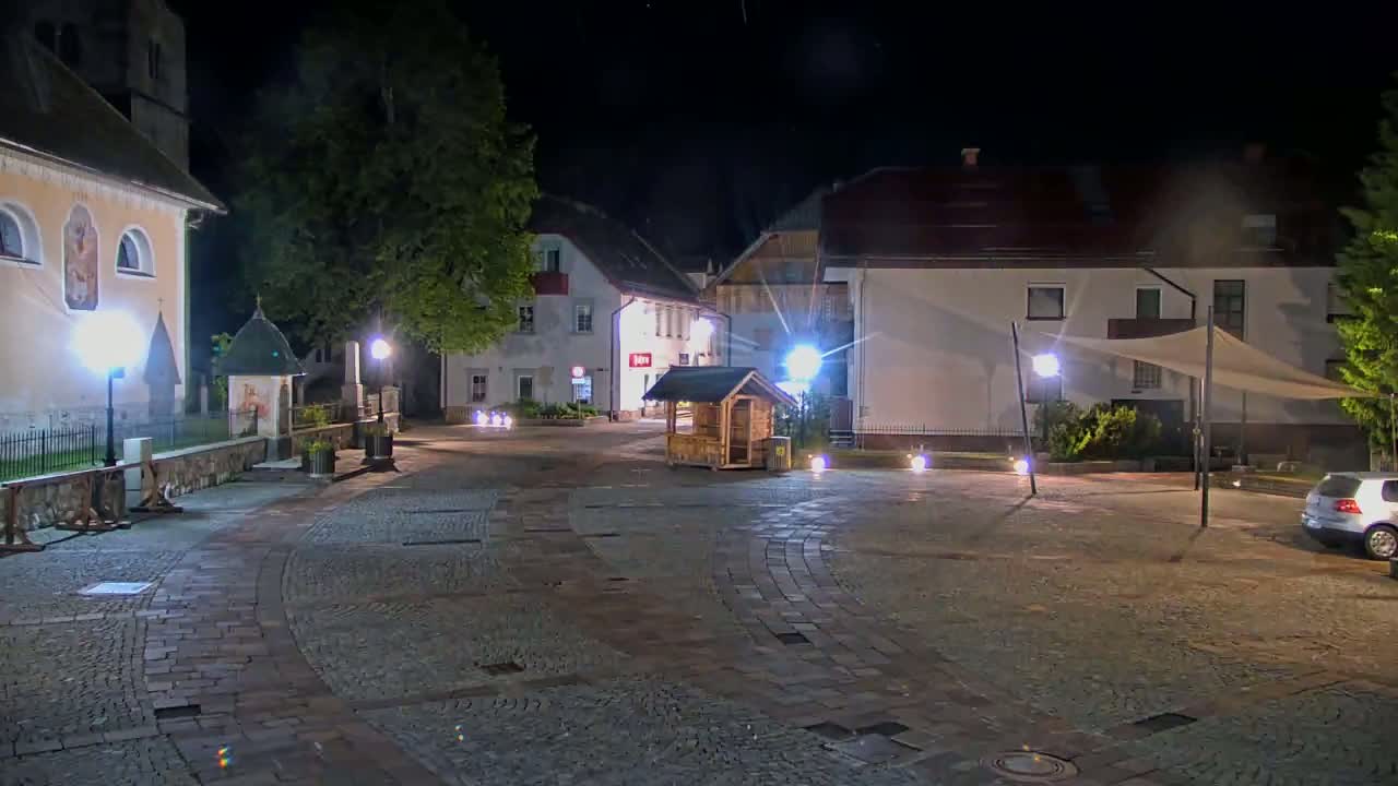 A nighttime view of a cobblestone town square, illuminated by streetlights, with buildings lining the perimeter and a small wooden structure in the center.