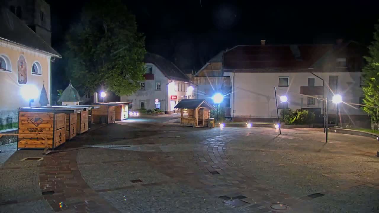 A nighttime view of a town square with several wooden stalls, buildings, and streetlights illuminating a paved area.