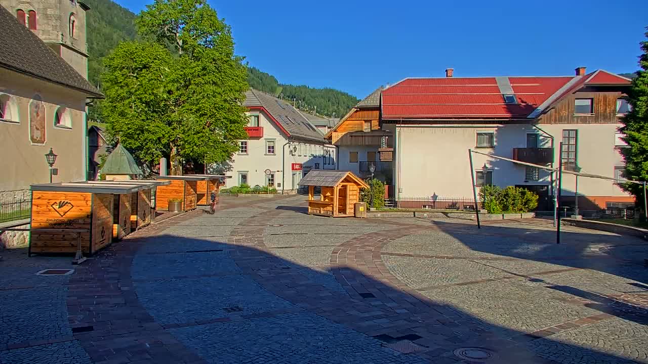 A sunny village square with several wooden stalls, buildings, and a volleyball net, is nestled in a valley against a backdrop of mountains.
