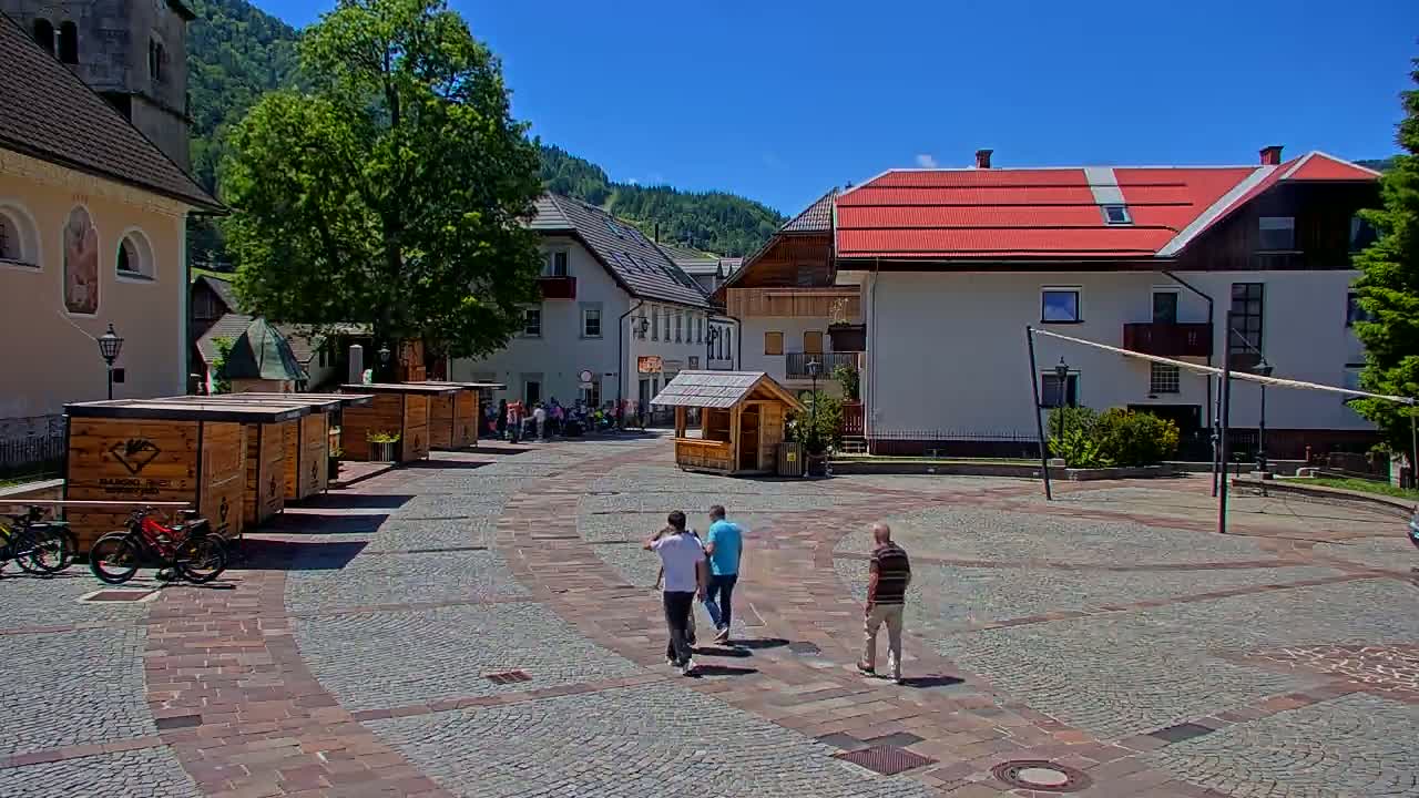 A cobblestone town square, under sunny skies, features several wooden kiosks, buildings, and a few people walking.