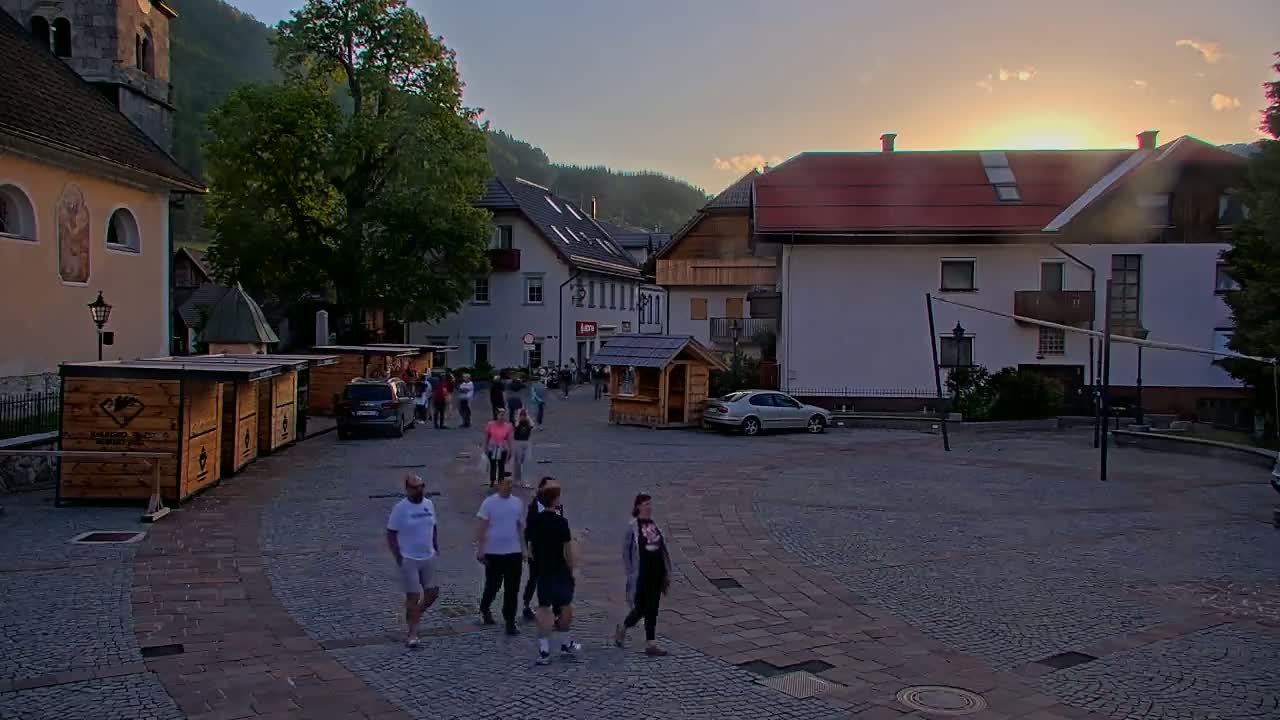 A cobblestone town square, surrounded by buildings and trees, with a few people walking around at sunset under partly cloudy skies.