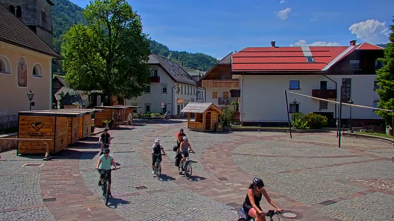 Several cyclists ride across a cobblestone town square on a sunny day, past wooden kiosks and buildings nestled in a mountain valley.