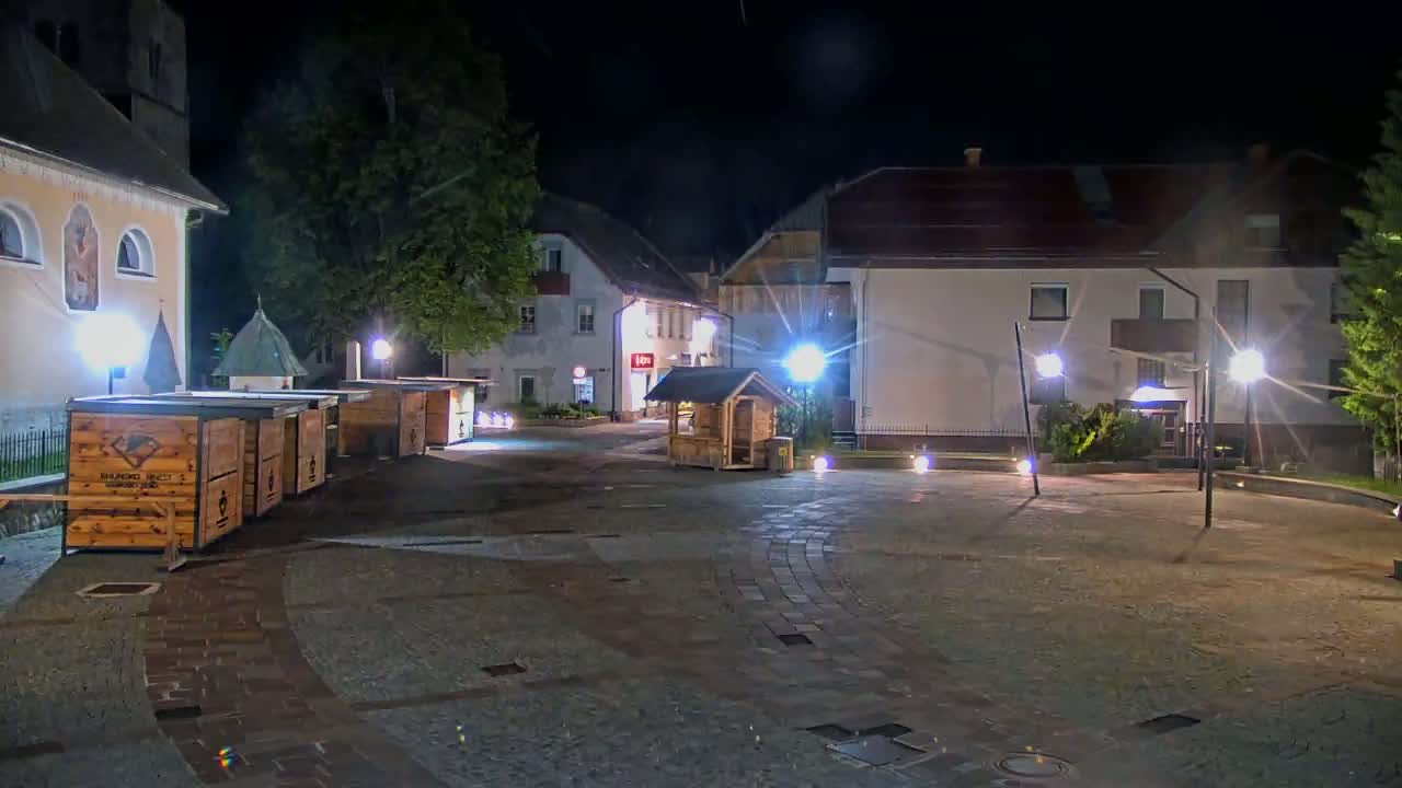 A nighttime view of a town square with several small wooden kiosks, buildings, and streetlights.