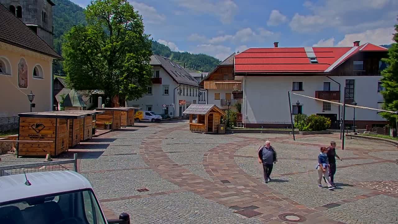 A paved town square with several buildings, wooden kiosks, and a few people walking on a sunny day.
