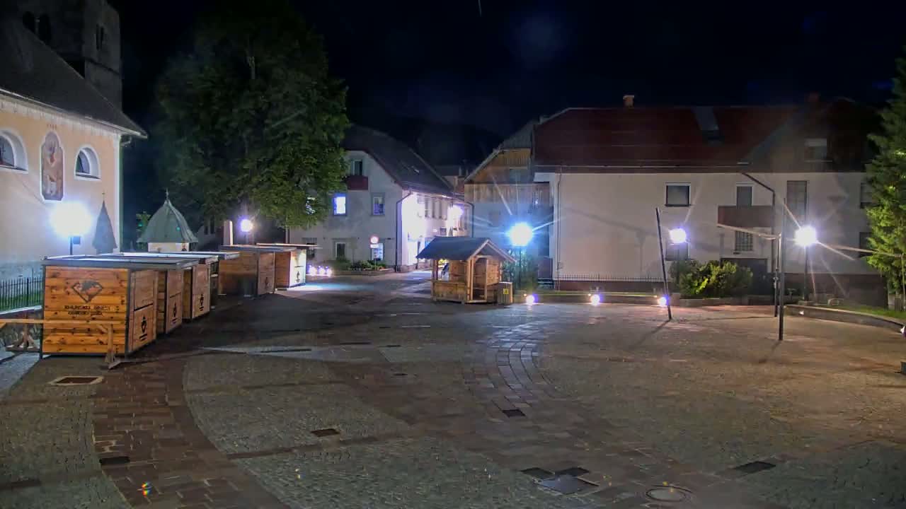 A nighttime view of a town square with several wooden stalls, buildings, and streetlights illuminating a paved area.