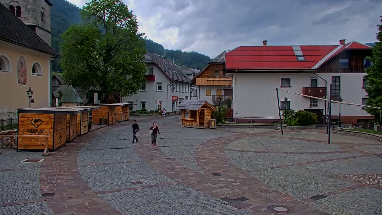 A paved town square with several small wooden kiosks, a few buildings, and two pedestrians, all under a cloudy sky.