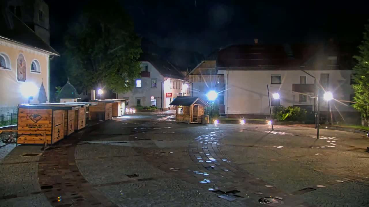A nighttime view of a town square with several wooden stalls, buildings, and streetlights illuminating a wet cobblestone surface.