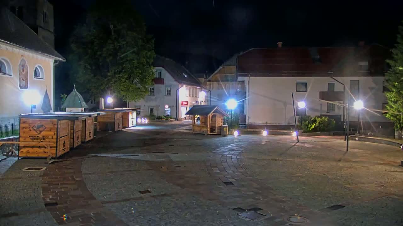 A nighttime view of a town square with several wooden stalls, buildings, and streetlights illuminating a paved area.