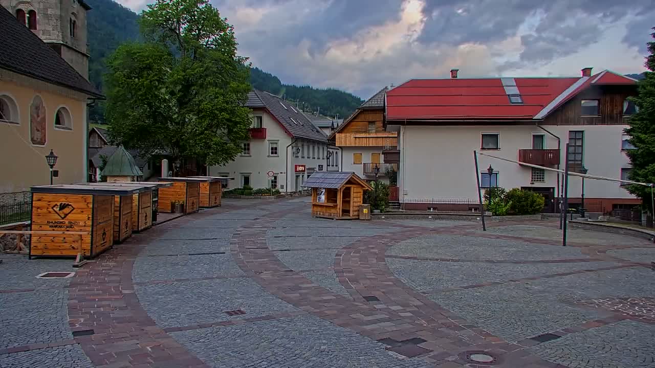 A cobblestone town square with several small wooden kiosks and buildings, under a partly cloudy sky.