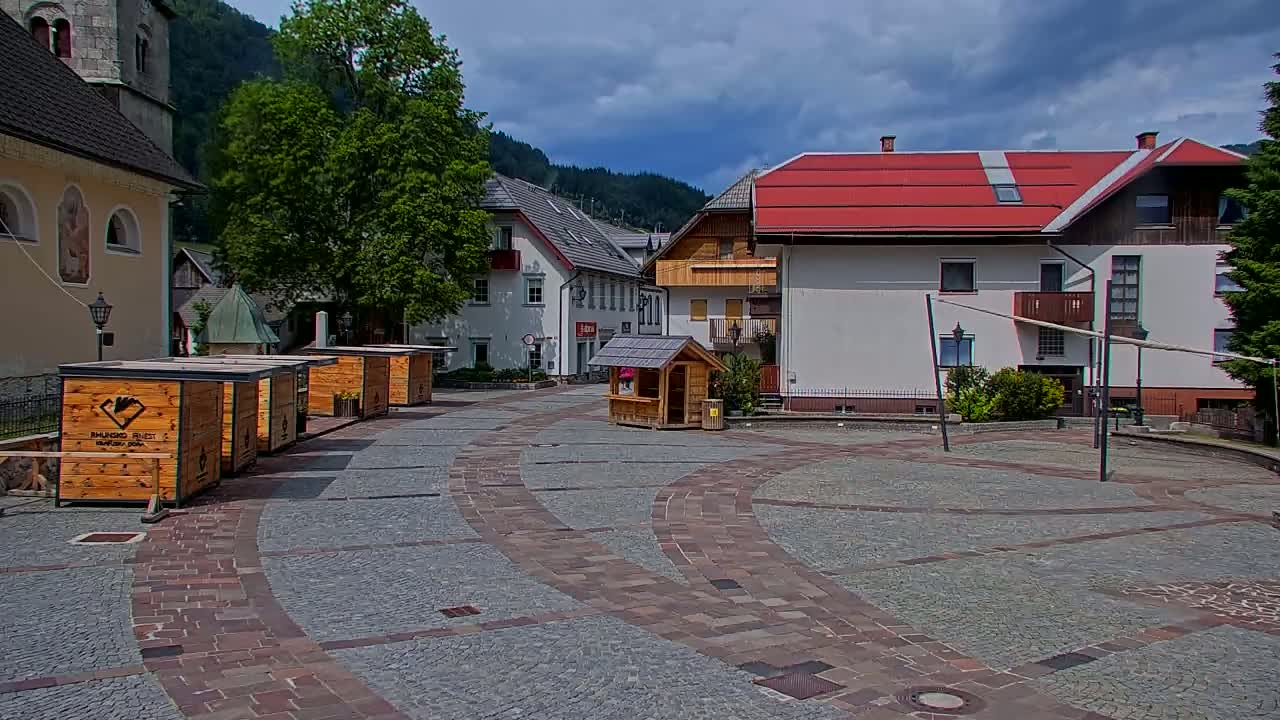 A cobblestone town square, under a partly cloudy sky, is surrounded by buildings, including a church and several small wooden kiosks.