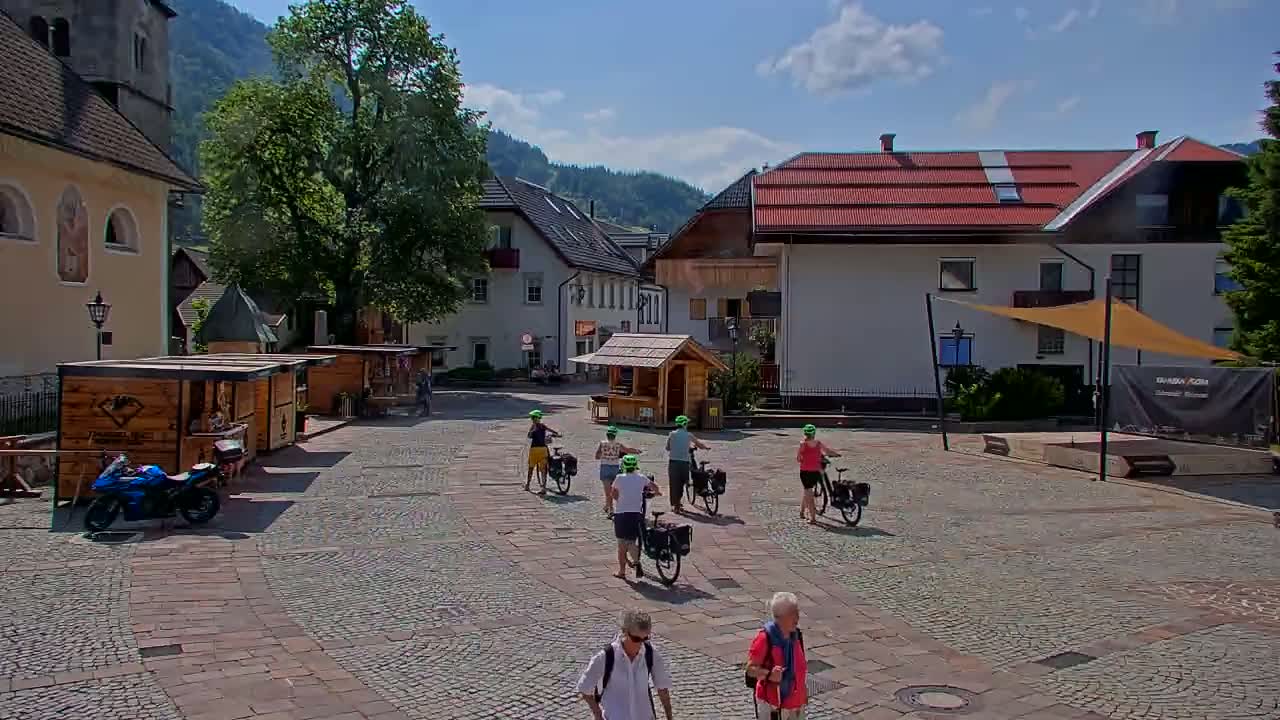A cobblestone town square, under sunny skies, features several people walking and riding bicycles past small wooden kiosks and buildings, including a church.