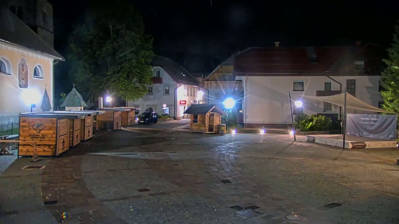 At night, a town square is illuminated, showing several wooden stalls, buildings, and a small stage under a canopy.