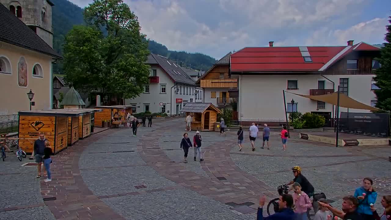 A cobblestone town square, surrounded by buildings and trees, is populated with pedestrians and cyclists under a mostly cloudy sky.