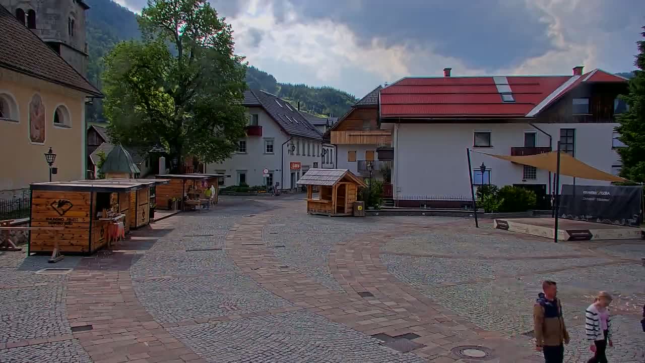 A cobblestone town square, under a partly cloudy sky, features several small wooden stalls, buildings with red roofs, and a few people walking.