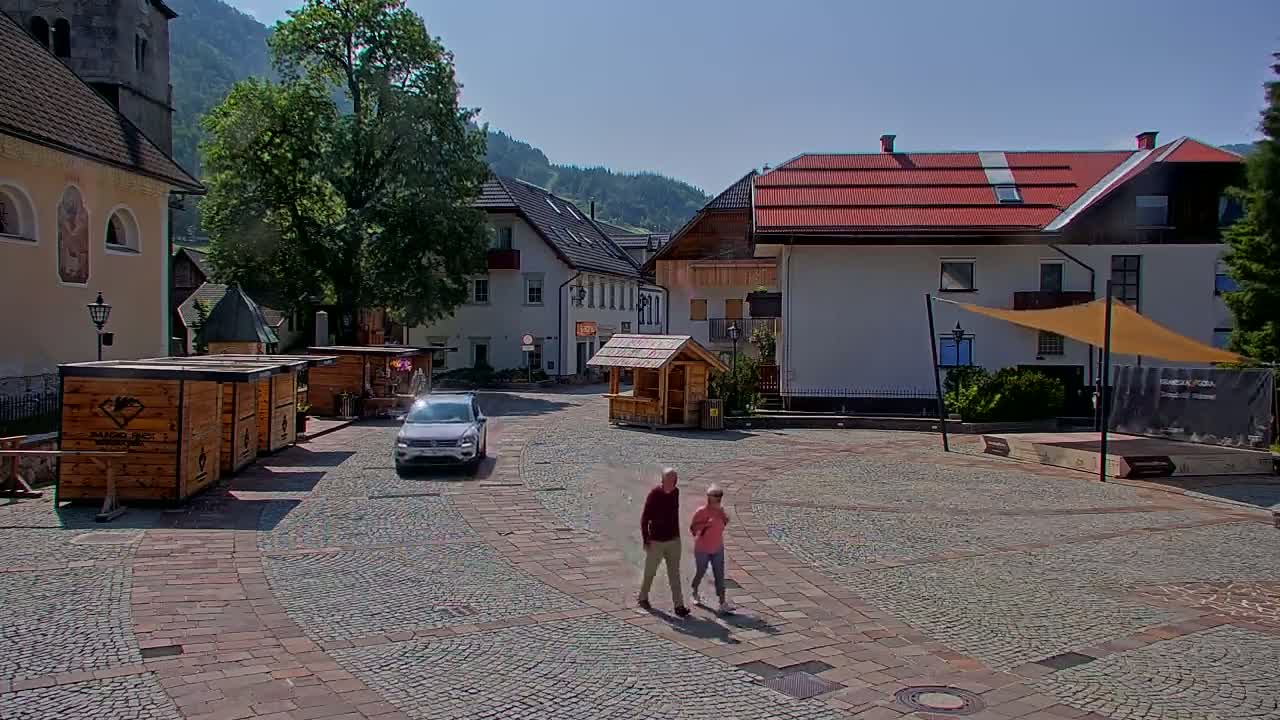 A paved town square with several buildings, wooden kiosks, and a car, under a sunny sky, features an elderly couple strolling across the cobblestones.