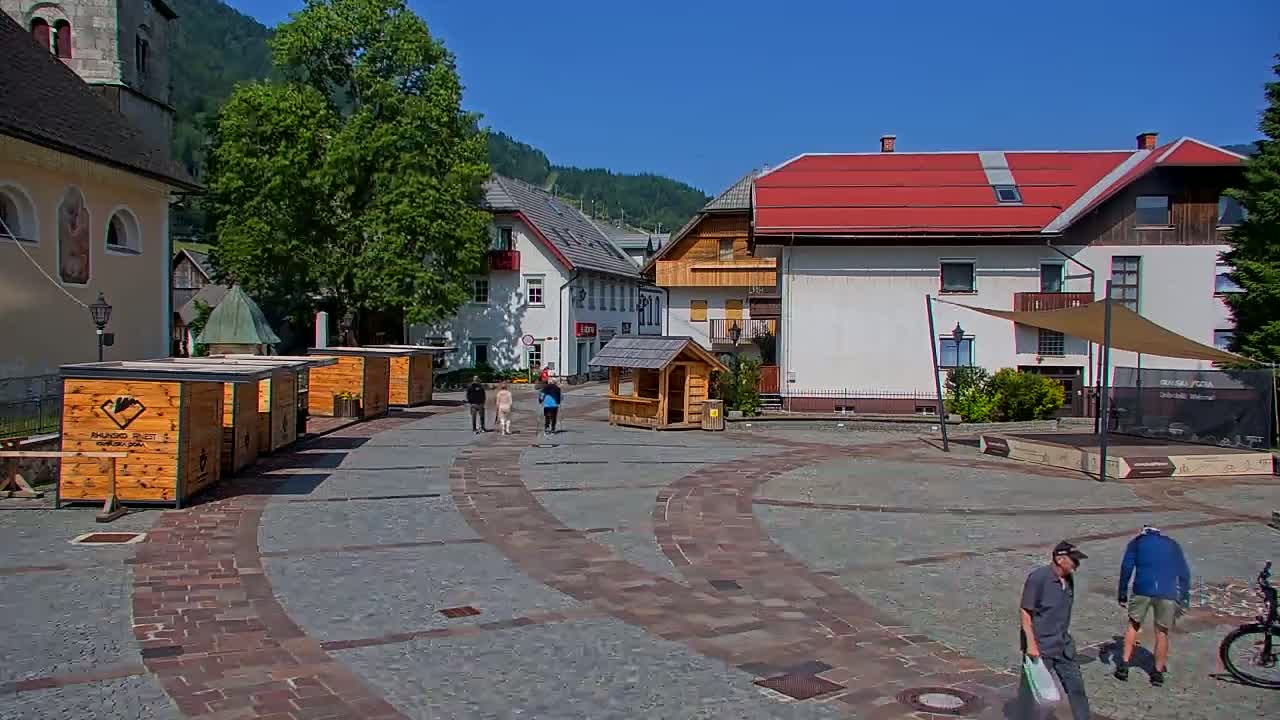 A paved town square with several small wooden kiosks, buildings with red roofs, and a few people walking on a sunny day.