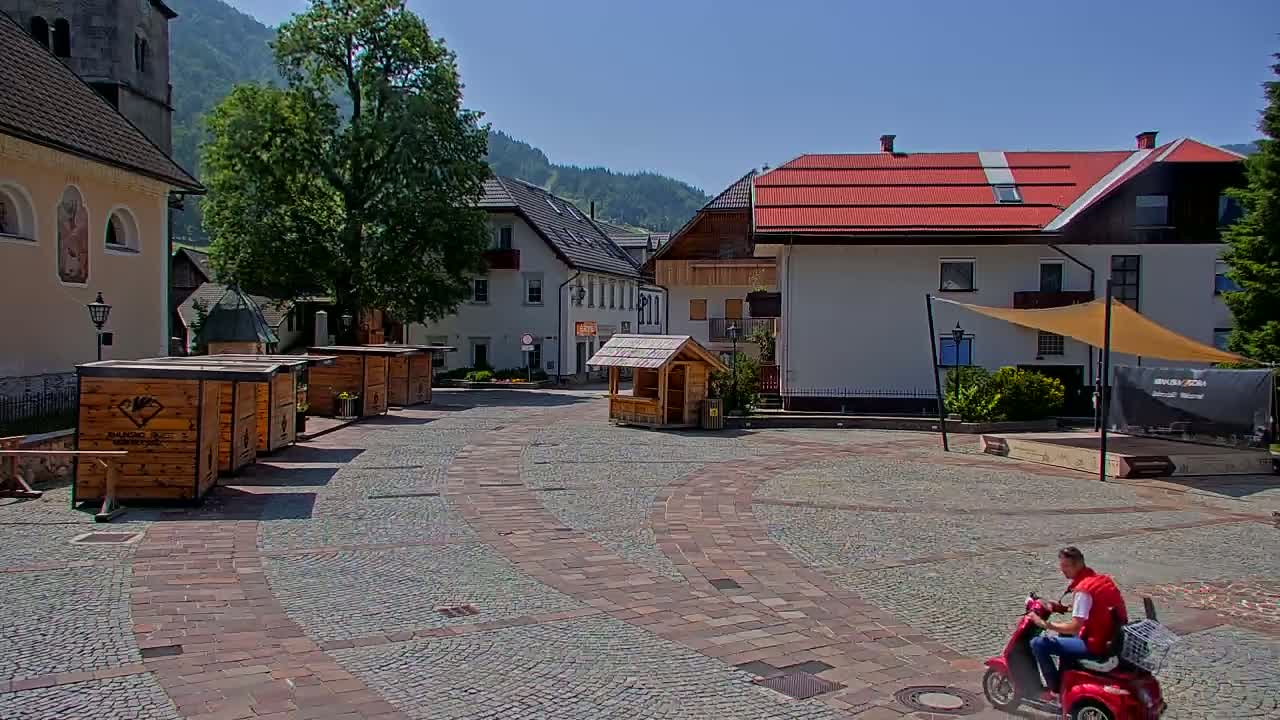 A man on a red scooter rides across a cobblestone town square on a sunny day, with several small wooden kiosks and buildings lining the square, set against a backdrop of mountains.