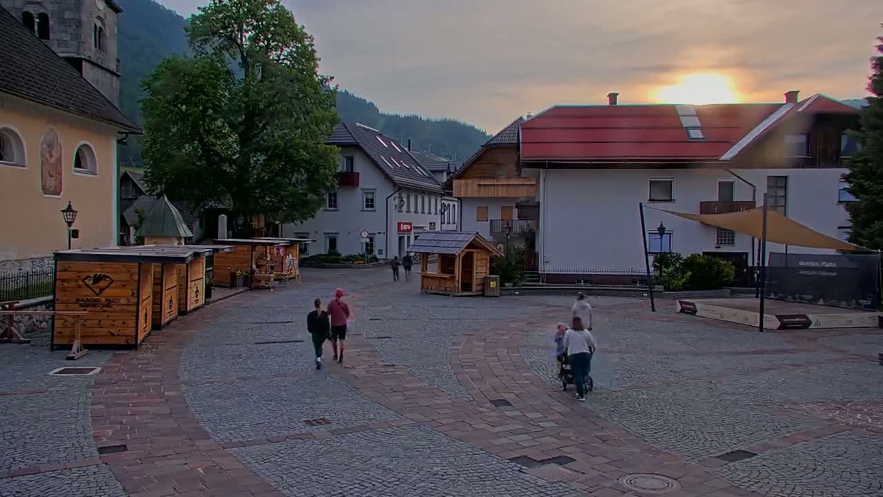 A cobblestone town square with several buildings, wooden stalls, and a few people walking around under a setting sun.