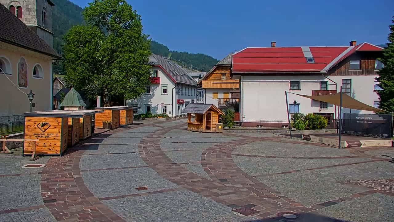 A paved town square with several small wooden stalls, buildings with red roofs, and a church on a sunny day.