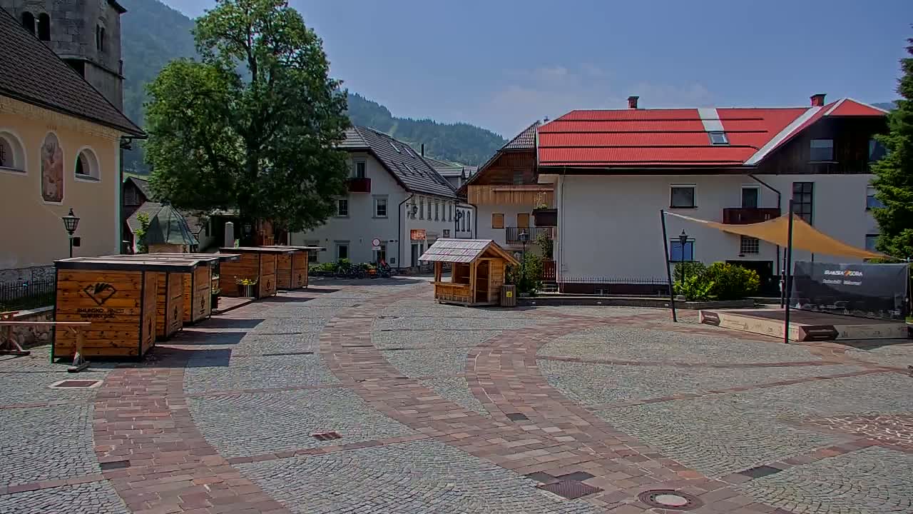 A town square with several small wooden kiosks, buildings with red roofs, and a cobblestone pavement under a clear, sunny sky.