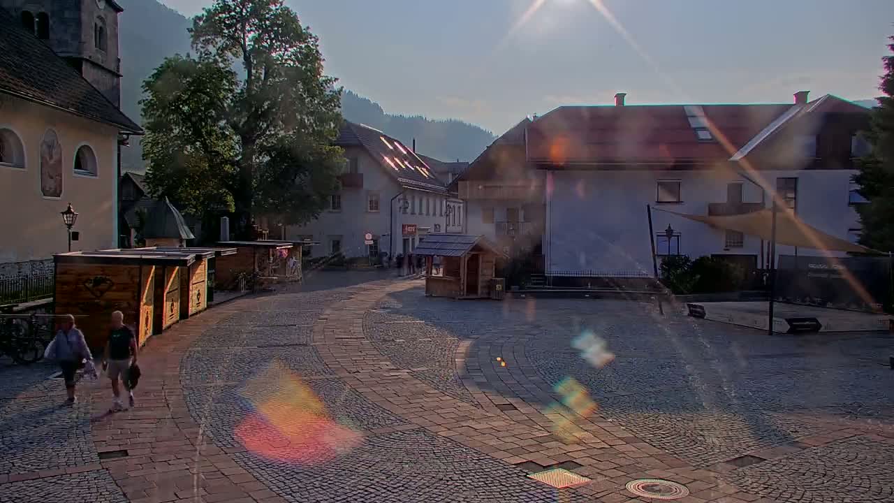 A cobblestone town square, partly shaded, with several small wooden kiosks and buildings, under a sunny sky.