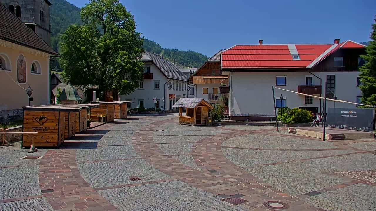 A cobblestone town square with several small wooden kiosks and buildings, under a clear sunny sky, is nestled against a backdrop of green hills.