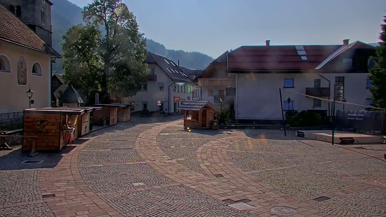 A cobblestone town square with several small wooden kiosks and buildings, under a sunny sky.