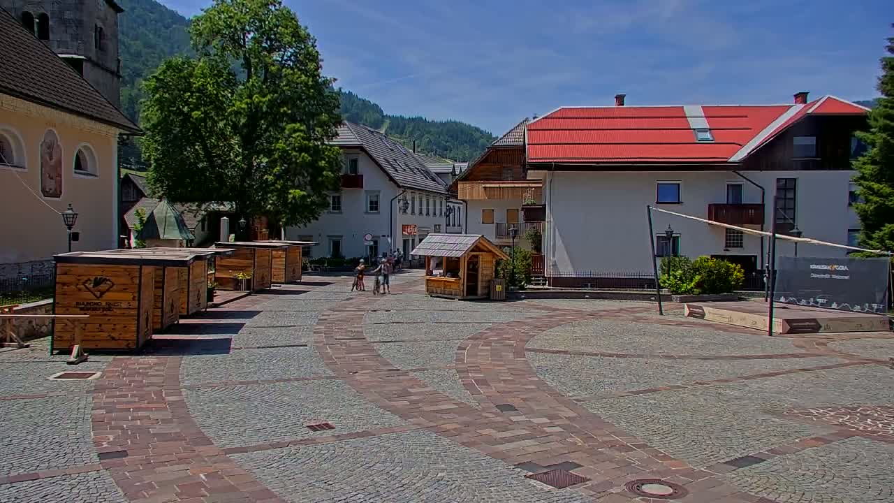 A paved town square with several small wooden kiosks, buildings with red roofs, and a few people, all under a sunny sky.