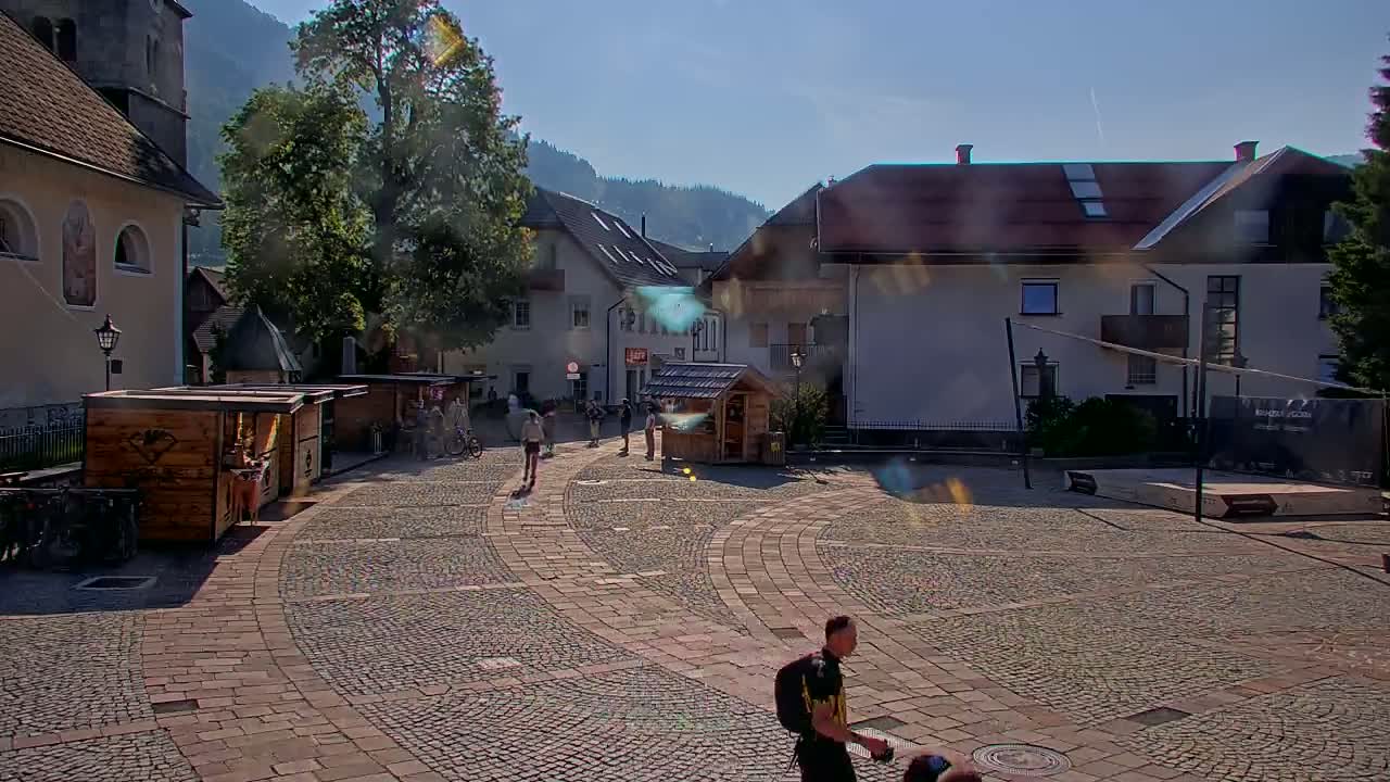 A cobblestone town square with several small wooden stalls, buildings, and a few people, all under a sunny sky.