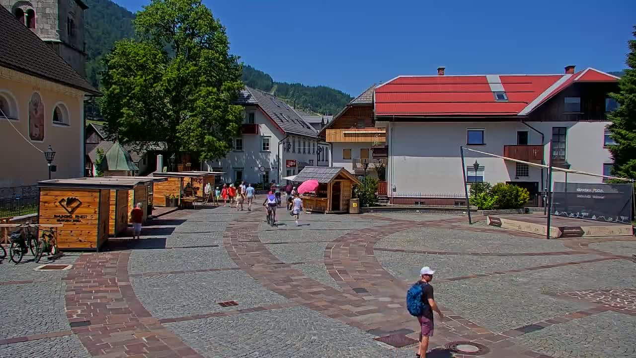 A cobblestone town square with several small wooden market stalls, buildings, and a few people walking on a sunny day.