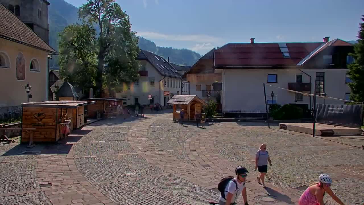 A cobblestone town square in a mountainous area on a sunny day features several small wooden stalls, buildings, and a few people walking and bicycling.