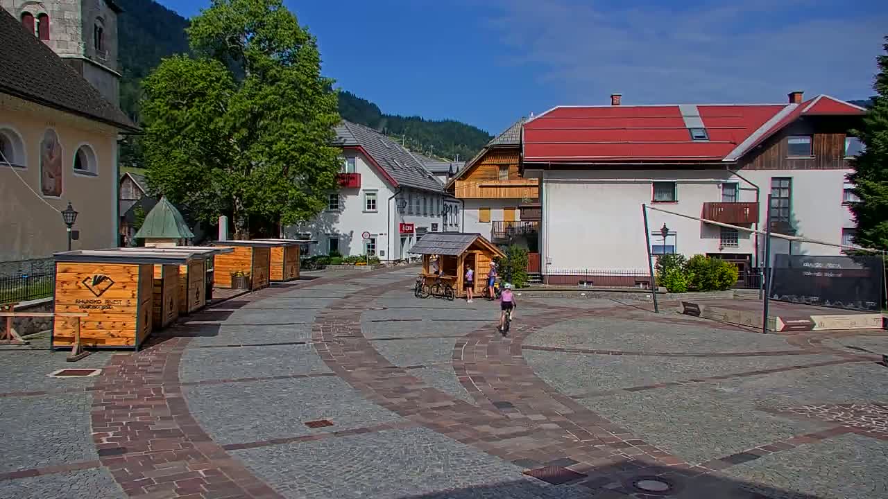 A sunny town square with several wooden kiosks, buildings, and a few people riding bicycles on a stone-paved area.