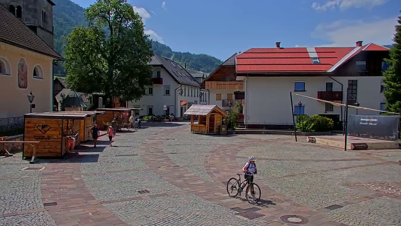 A cyclist rides across a cobblestone town square, surrounded by buildings, small wooden kiosks, and a background of mountains under a sunny sky.