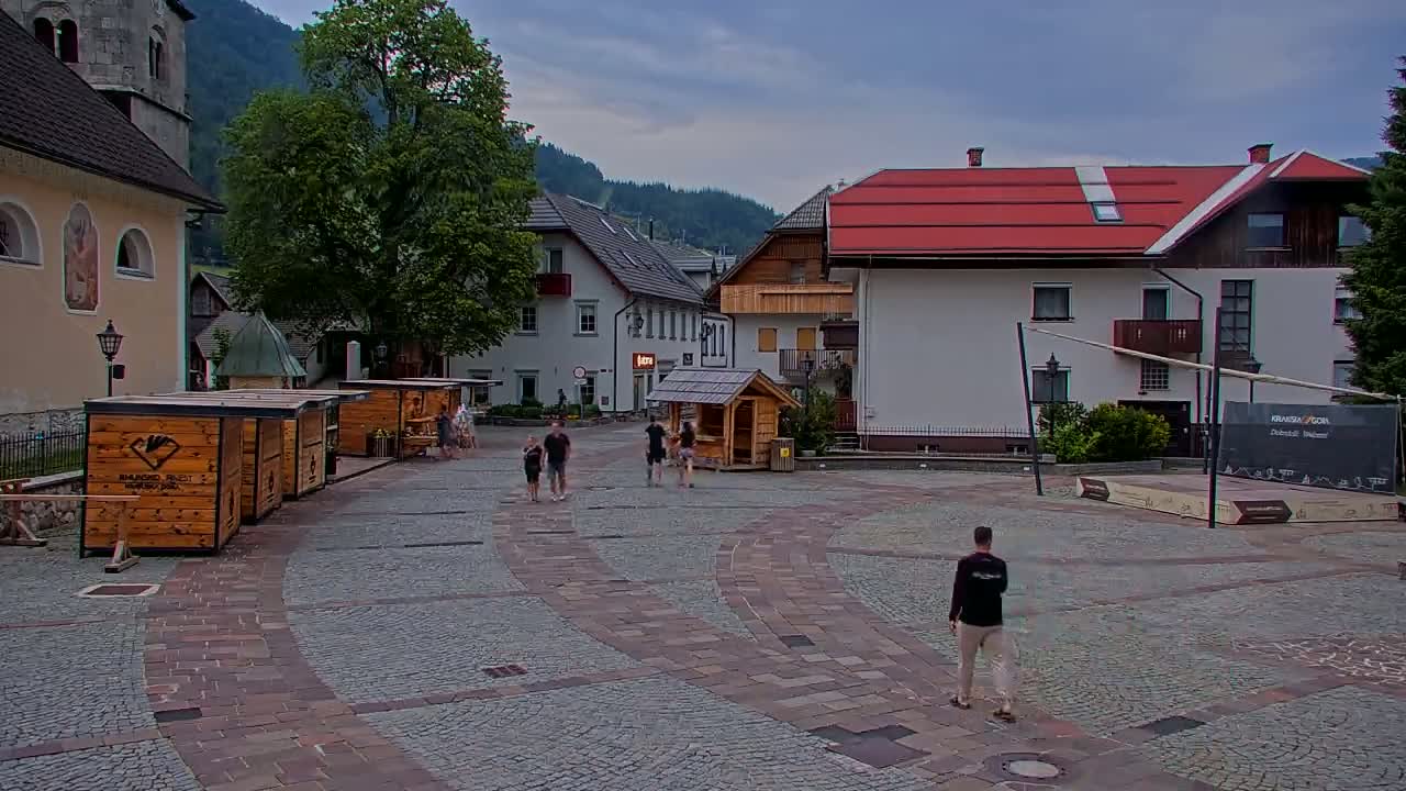 A cobblestone town square with several small wooden kiosks, buildings with red roofs, and a few people walking under a partly cloudy sky.