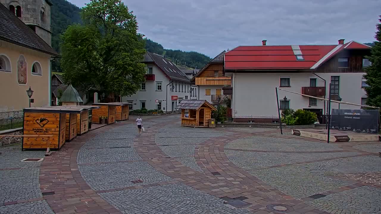 A person walks across a cobblestone town square with several small wooden kiosks and buildings, under a cloudy sky in a mountainous region.