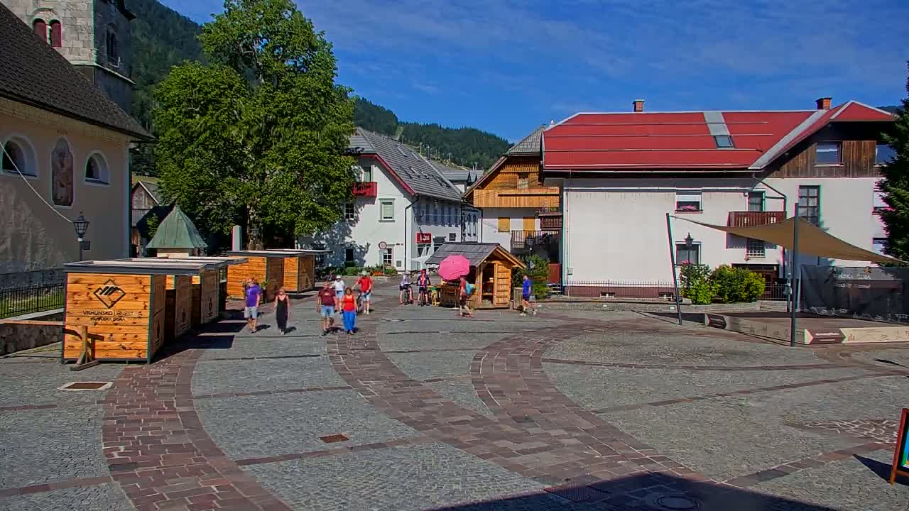 A sunny day in a village square shows several people walking past wooden kiosks and buildings, with mountains in the background.