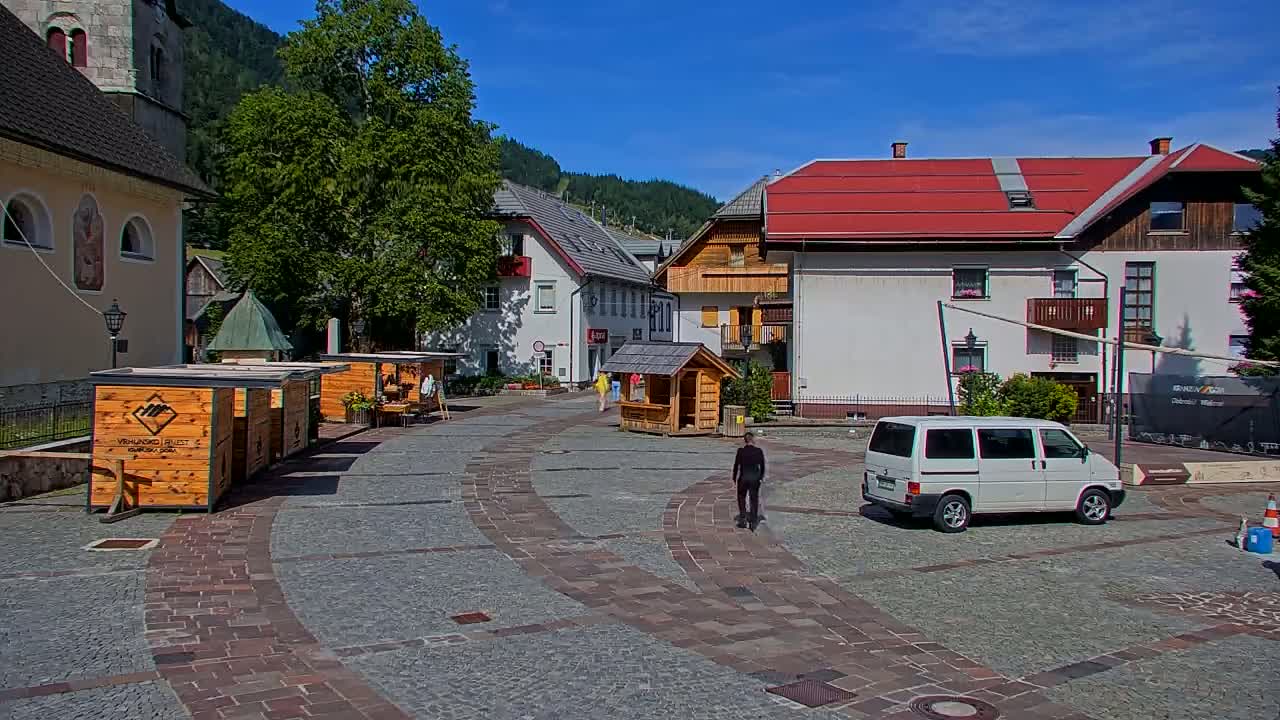 A paved town square with several buildings, small wooden kiosks, and a white van is visible on a sunny day, with mountains in the background.