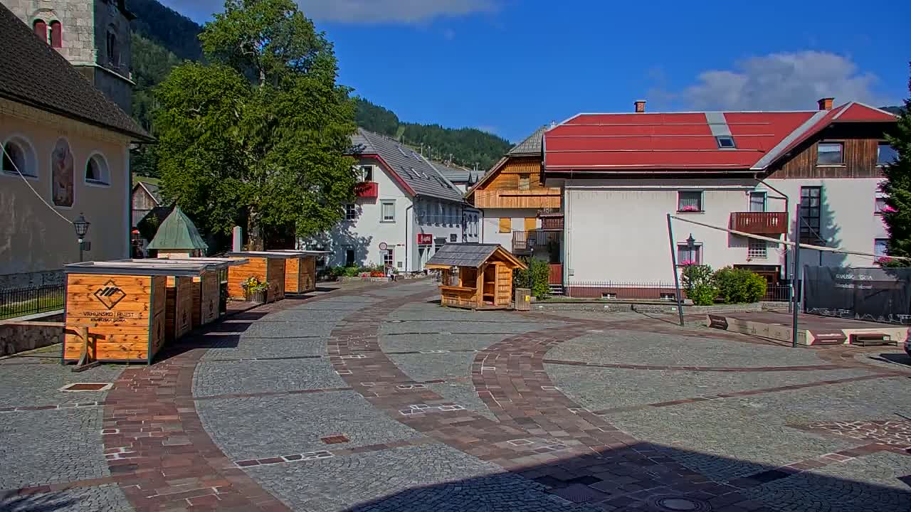A town square with wooden market stalls and buildings, under a sunny sky, is surrounded by mountains.