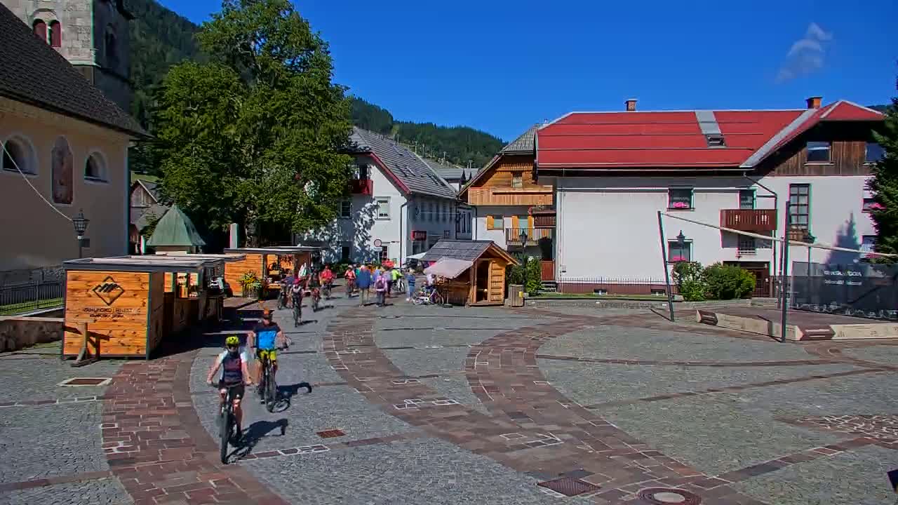 A paved town square in a mountain village on a sunny day features several cyclists and pedestrians, wooden market stalls, and buildings with red roofs.