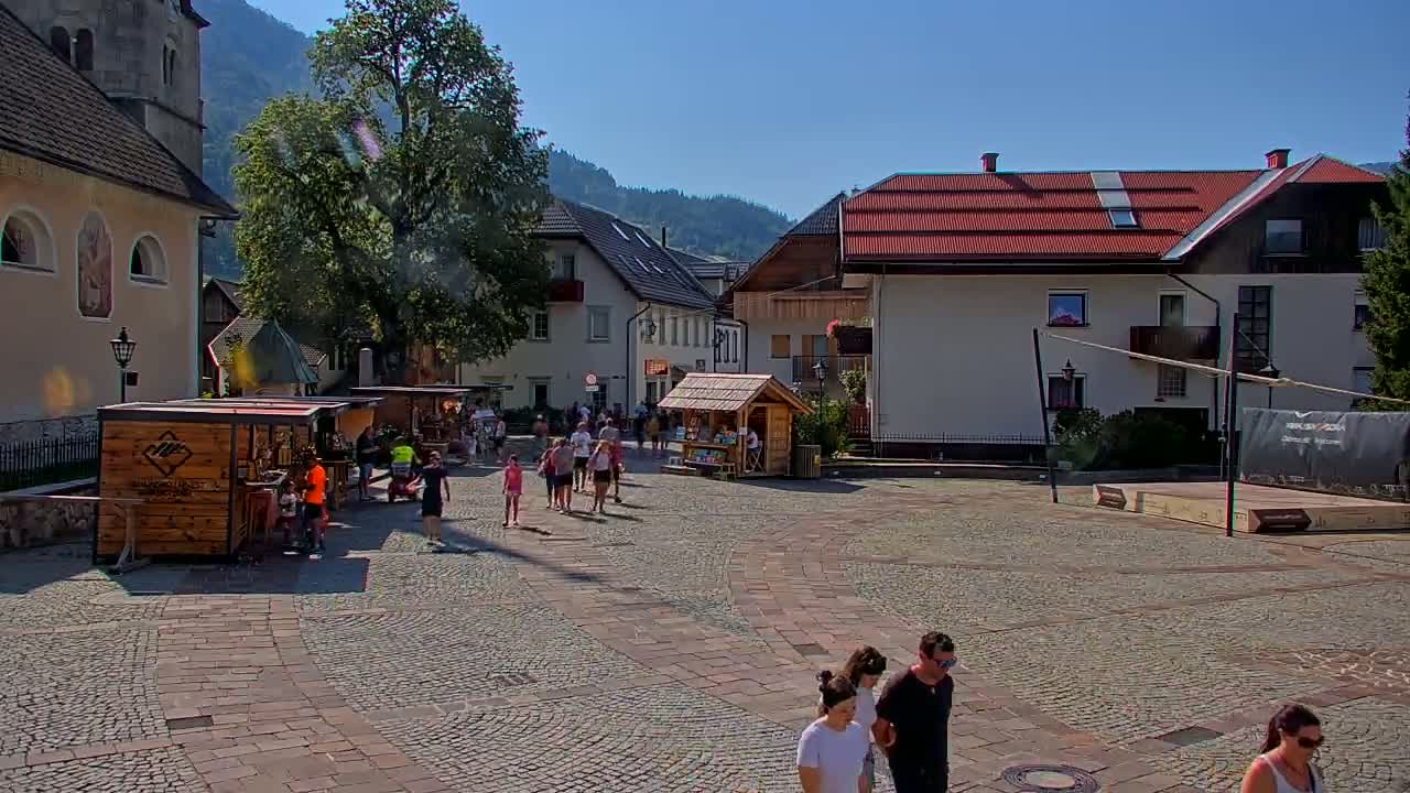 A paved town square in a mountain village on a sunny day features several cyclists and pedestrians, wooden market stalls, and buildings with red roofs.