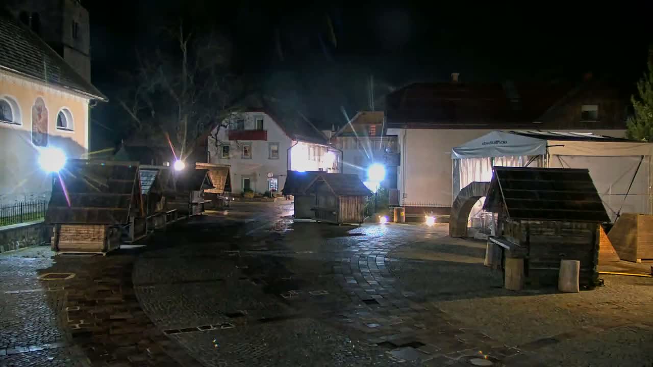 A nocturnal view of a wet, empty town square filled with wooden market stalls and illuminated by artificial light, flanked by buildings including a church and a large white tent, under a clear night sky.