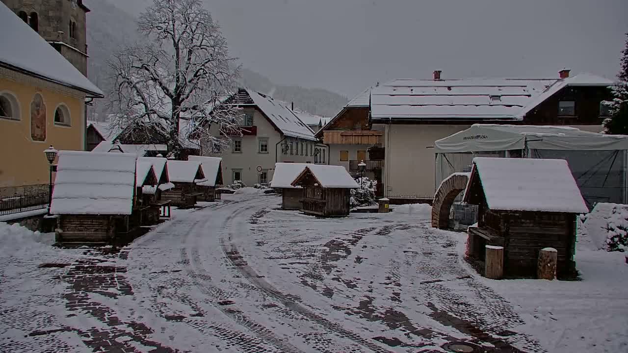 A quaint village square is completely blanketed in fresh snow, with several charming wooden stalls, residential buildings, and a church tower visible under a grey, overcast winter sky.