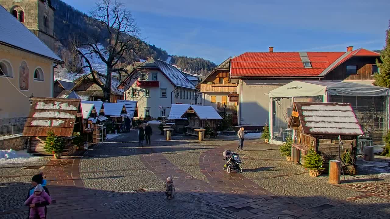 A sunny winter day illuminates a quaint village square featuring market stalls with snow-dusted roofs, several people, and distant snow-capped mountains under a clear blue sky.